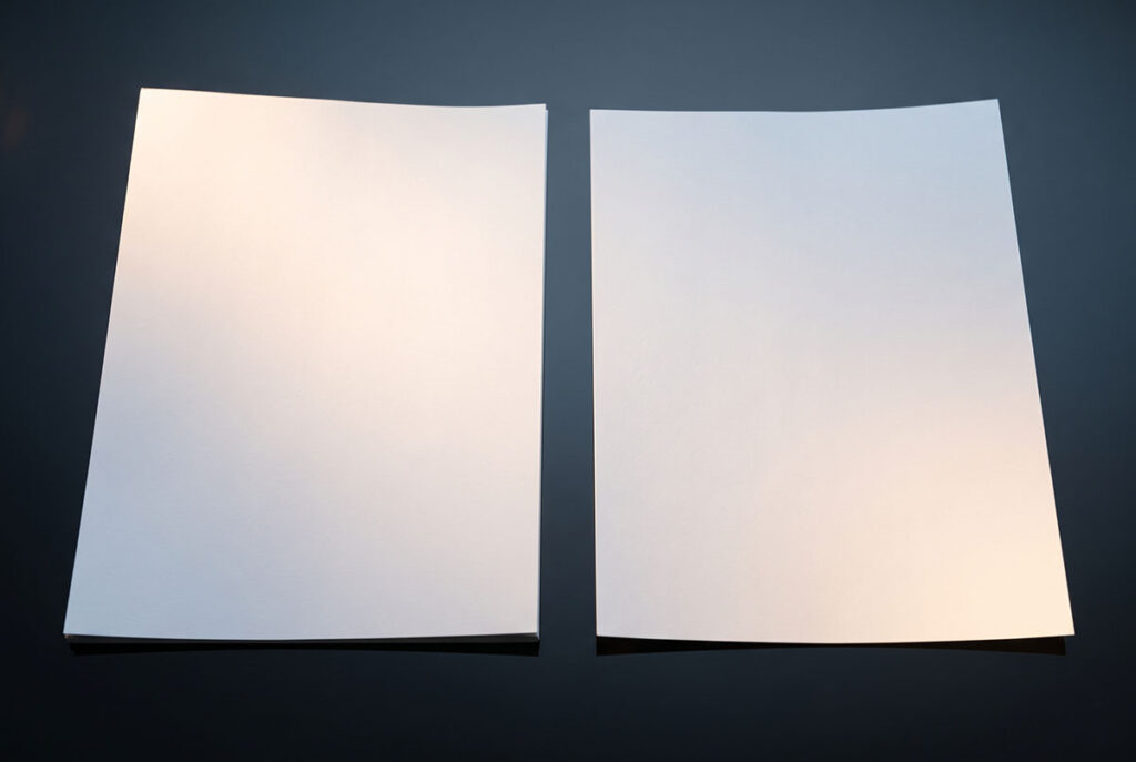 Two near-identical documents on a dark reflective surface under controlled lighting, suggesting the collapse of proof of authenticity in digital media.
